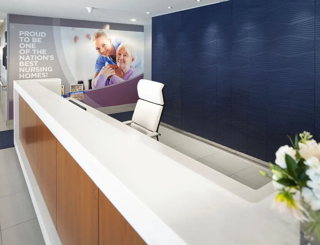 A modern reception area with a white countertop and a white office chair. The back wall features a large poster of a caregiver smiling at an elderly woman, with the text 