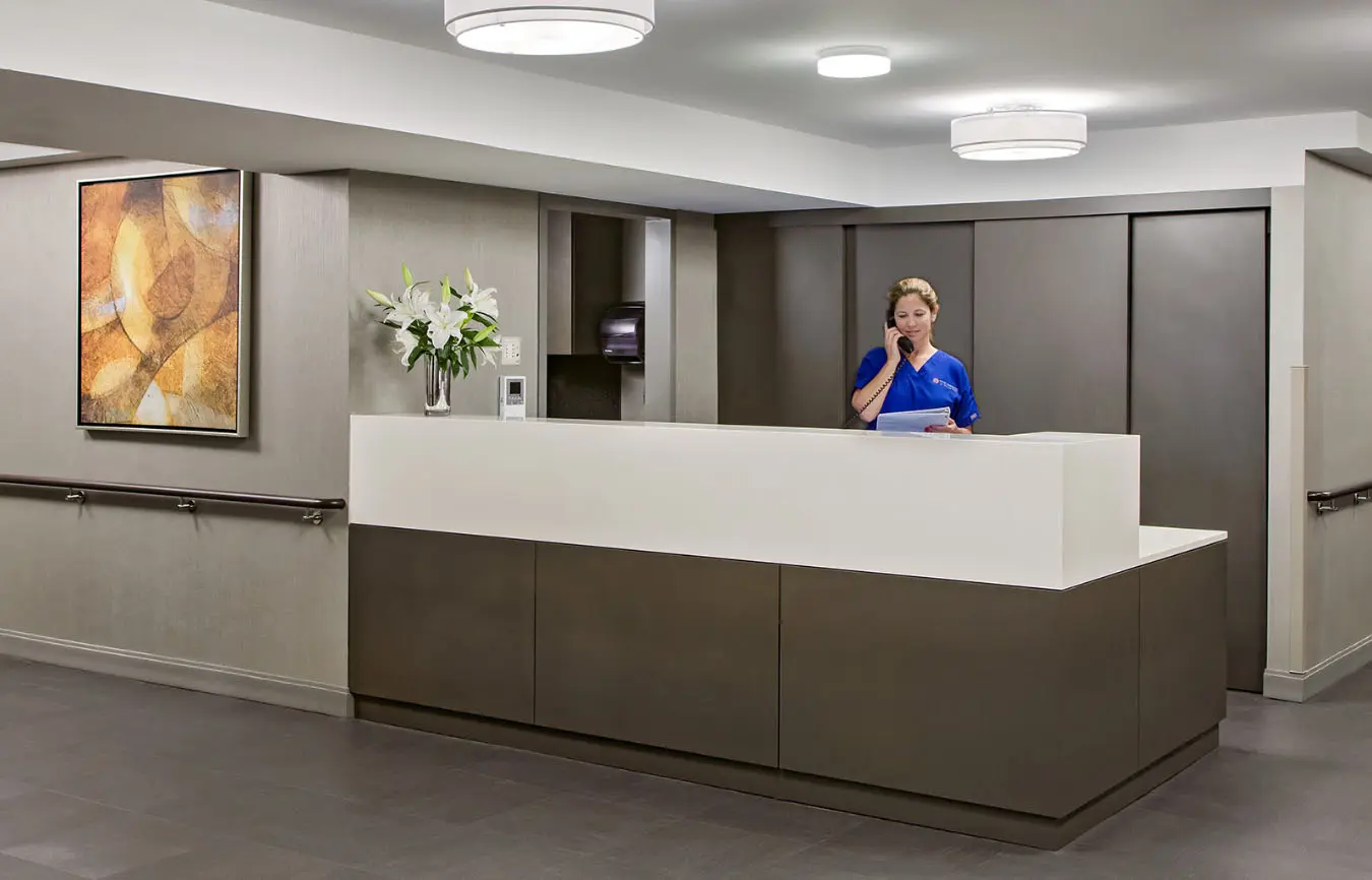 A receptionist in a blue uniform standing behind a white and brown reception desk, talking on the phone and holding some papers in a modern, well-lit hospital or office lobby.